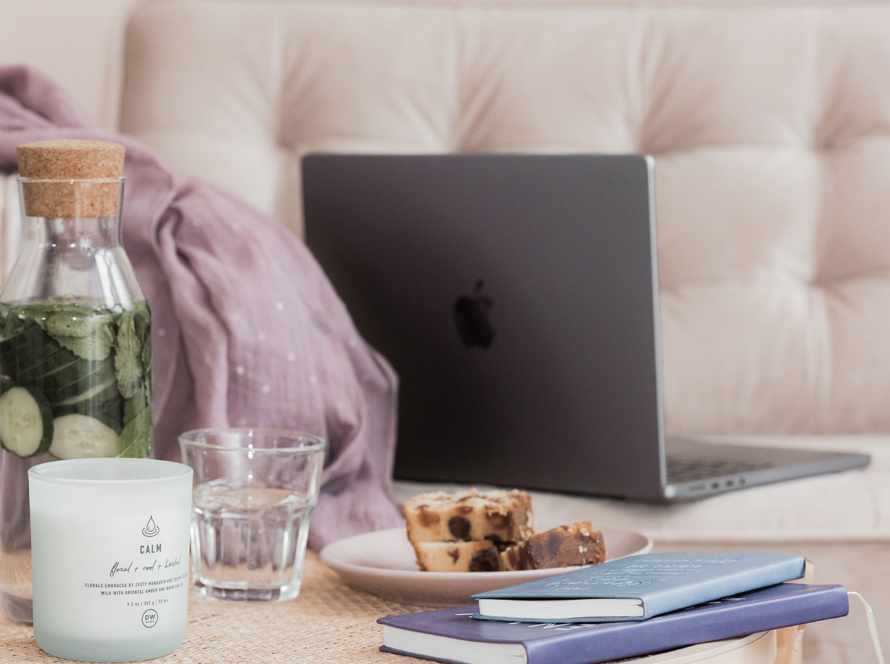 Laptop on sofa with notebooks, candle, glass water, cake and more.