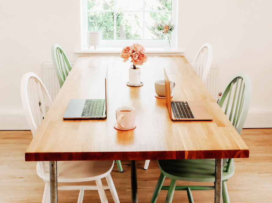 Wooden table with white and green wooden chairs around it. Laptops on the table and a view of green leaves out of the window.