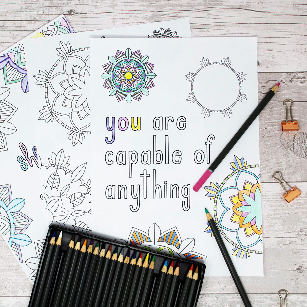 Image of partially coloured colouring sheets on a pale wood background with coloured pencils and mini bulldog clips.