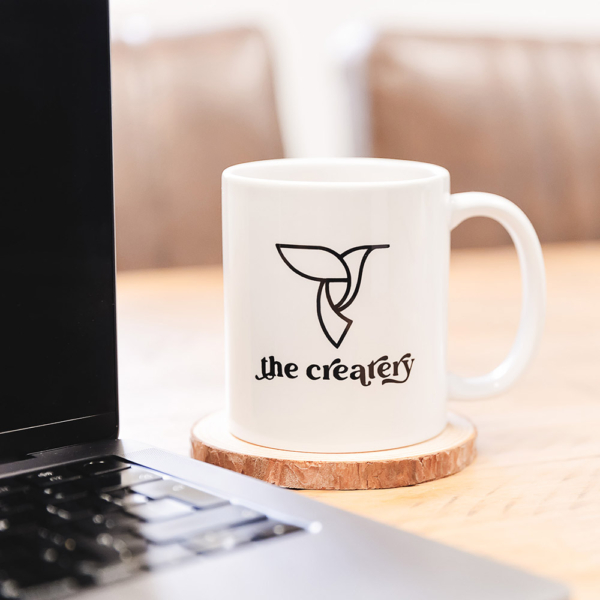 White mug with the Createry logo, and a laptop on a wooden desk.