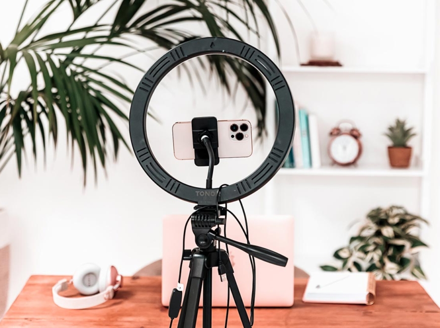Ring light on wooden desk with plant and white ladder bookshelf against white background.